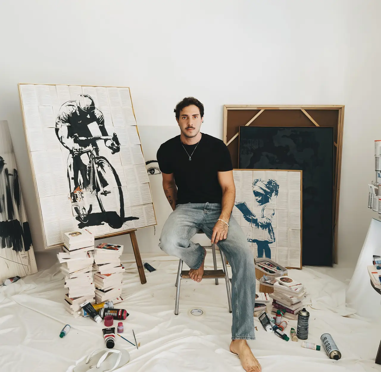 O artista João Pedro Biolchini sentado no ateliê entre quadros pintados sobre páginas de livro
