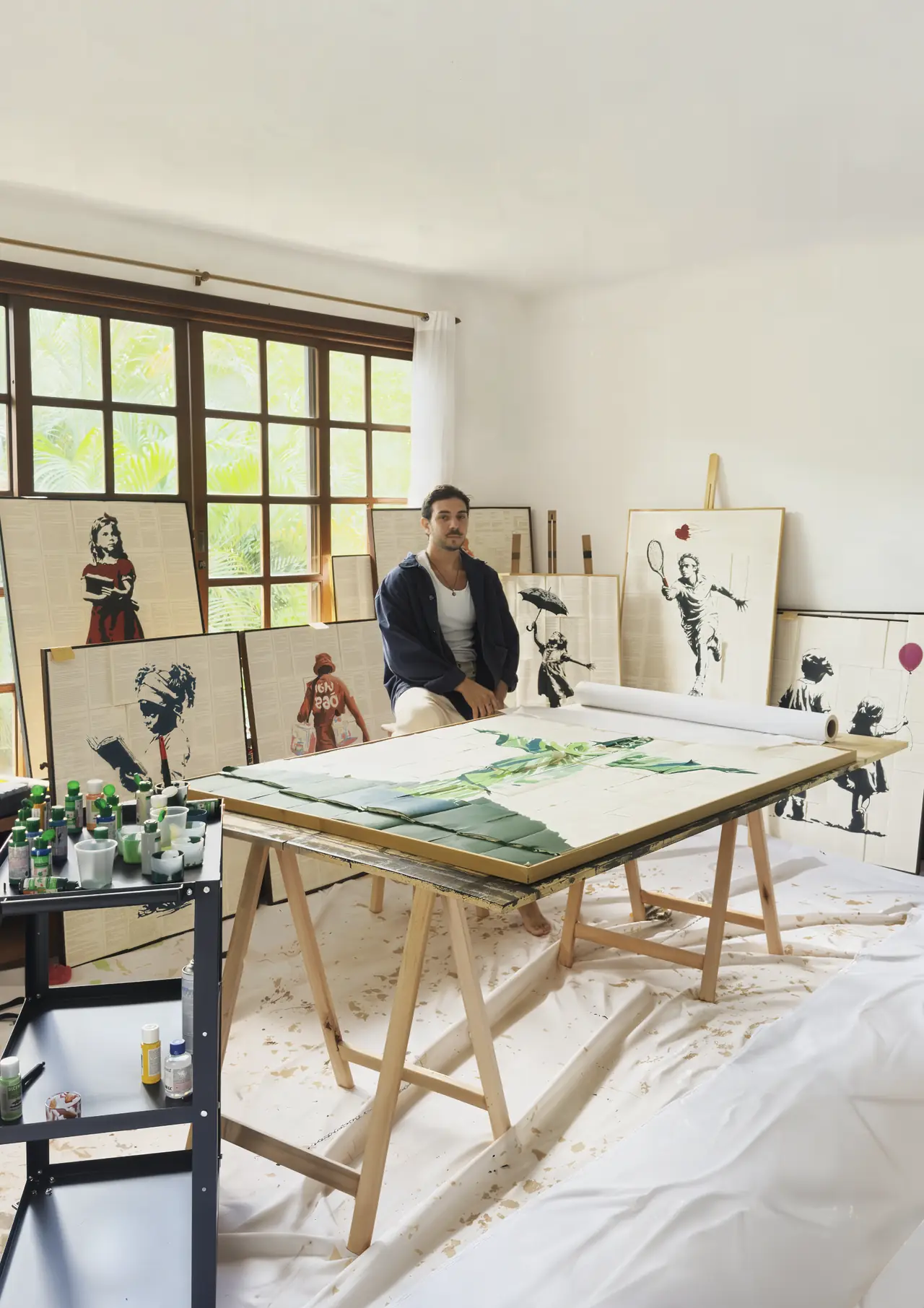 Ateliê DoJoão com várias obras pintadas sobre páginas de livro encostadas na parede e uma obra verde em produção sobre a mesa